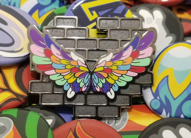 May include: A silver metal pin with a brick wall design and a colorful butterfly wing design. The wings are made up of multiple colors, including red, orange, yellow, green, blue, purple, and pink.
