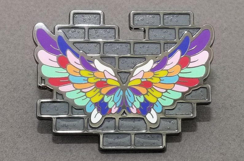 May include: A colorful enamel pin featuring a pair of wings breaking through a brick wall. The wings are made up of multiple colors, including purple, pink, red, orange, yellow, green, blue, and white.