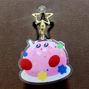 May include: A pink, cartoon-style keychain featuring a character with blue eyes, red cheeks, and a white horn. The keychain has a gold star and a clear gem at the top, with a gold chain. The character is on a white plate with colorful decorations.
