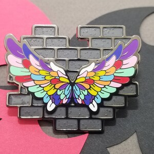 May include: A silver metal pin with a brick wall design and a pair of colorful wings. The wings are made up of many small feathers in shades of purple, pink, blue, green, yellow, orange, and red.