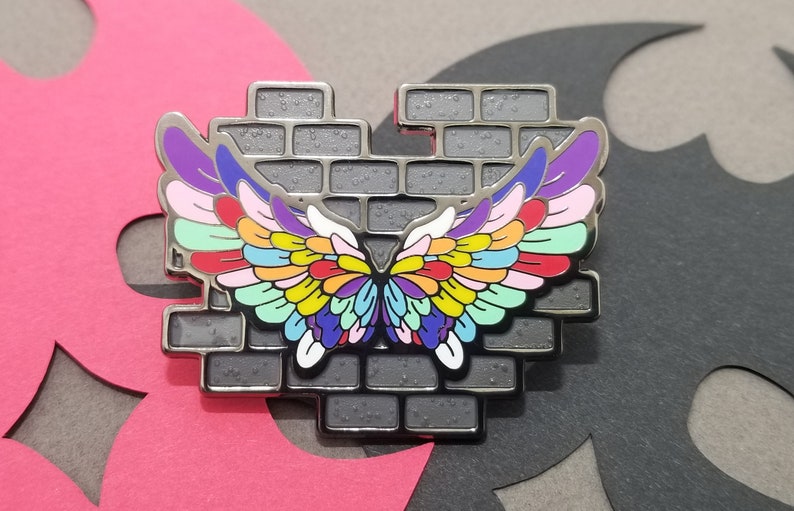May include: A silver metal pin with a brick wall design and a pair of colorful wings. The wings are made up of many small feathers in shades of purple, pink, blue, green, yellow, orange, and red.