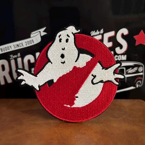 May include: Embroidered patch featuring the Ghostbusters logo. The patch has a red circle with a white ghost and a red slash through it. The ghost is white with black outlines and details. The patch is on a brown surface.