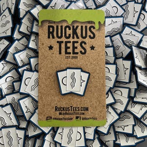 Puede incluir: Un pin que presenta tres tarjetas blancas con arte lineal negro de una línea ondulada, un semicírculo y una estrella. El pin está sobre una tabla de corcho con el texto "RUCKUS TEES EST. 2009". El fondo está lleno de pines similares.