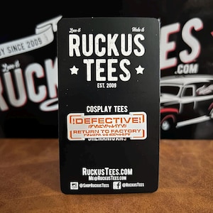 May include: Black business card with the text "Ruckus Tees" and "Est. 2009". The card also features the text "Cosplay Tees" and a sticker that says "Defective!" and "Return to Factory".