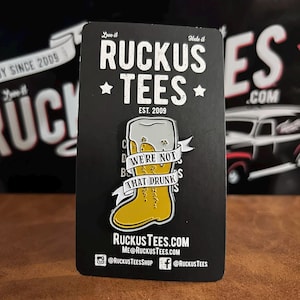 May include: A black enamel pin depicting a yellow beer boot with a foamy top and the text "WE'RE NOT THAT DRUNKS". The pin is on a black card with the text "RUCKUS TEES" and website details.