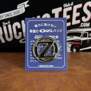 Könnte beinhalten: Ein silberfarbener Metall-Anstecker mit einem großen schwarzen "Z"-Design, auf einer blauen Karte mit japanischem Text. Der Anstecker ist kreisförmig mit einem erhabenen Rand. Die Karte hat zusätzlichen Text und die Website-Adresse RUCKUSTEES.COM.