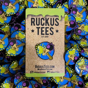 May include: A collection of enamel pins featuring a cartoon mushroom design with a blue, green, and brown color scheme. The pins are on a corkboard backing with the text "RUCKUS TEES" and "EST. 2009". The pins are scattered across a dark surface.