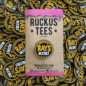 May include: A collection of black and yellow circular pins with the text "CERTIFIED RAY'S OCCULT N.Y.C." and a corkboard with the text "RUCKUS TEES EST. 2009". The corkboard has a pink border with the text "Love it. Hate it."
