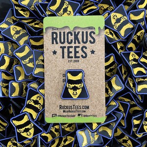 May include: A black and yellow enamel pin featuring a masked face design, set on a corkboard card with the text "Ruckus Tees" and website information. The pin is surrounded by similar pins.