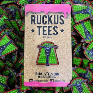 May include: A pin featuring a green and black design with a purple and red outline, mounted on a corkboard card. The card has the text "RUCKUS TEES EST. 2009" and social media handles. The pin is surrounded by other pins.