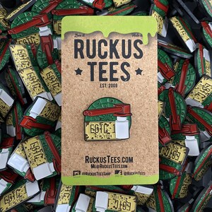 May include: A collection of enamel pins featuring a New York license plate design with the text "EOTC-1 NEW YORK" and a red and green vehicle. The pins are displayed on a corkboard with the "Ruckus Tees" logo and website information.