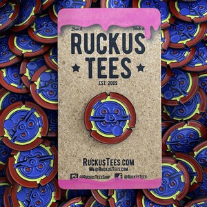 May include: A collection of enamel pins with a circular design. The pins feature a blue and yellow central design with a red outer ring. The pins are on a corkboard with the text "RUCKUS TEES" and website information.