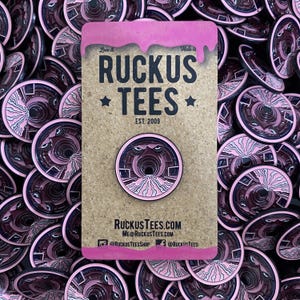 May include: A pink and black enamel pin featuring a tunnel design with the text "RUCKUS TEES EST. 2009". The pin is displayed on a backing card with the brand's website and social media handles. Many similar pins are scattered around the card.