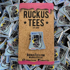 Puede incluir: Un pin con un diseño de mochila de protones negro y amarillo sobre un fondo azul y blanco. El pin está sobre un corcho con el texto "Ruckus Tees" e información del sitio web. Otros pines con el mismo diseño lo rodean.