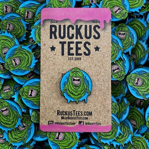 May include: A green enamel pin featuring a cartoon character with a wide grin, surrounded by a blue outline. The pin is mounted on a corkboard with the text "RUCKUS TEES EST. 2009". The background is filled with similar pins.