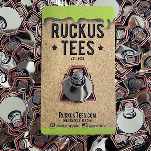 May include: A collection of silver-colored enamel pins with a paint tube design, outlined in red and blue. A corkboard card displays a pin with the text "RUCKUS TEES EST. 2009" and social media handles. The pins are scattered across the surface.