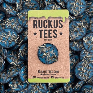 May include: A blue and gold enamel pin featuring the text "Tobin's Spirit Guide" inside a starburst design. The pin is displayed on a corkboard card with the "Ruckus Tees" logo and website address. The background is filled with similar pins.