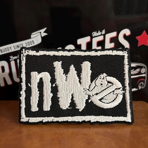 Könnte beinhalten: Schwarzer rechteckiger Aufnäher mit weiß gesticktem Text und Bildern. Der Text lautet "nWo" mit einem Ghostbusters-Logo auf der rechten Seite. Der Aufnäher hat einen abgenutzten Rand. Der Hintergrund enthält den Text "BUDDY SINCE 2009" und "Halo it TEES".