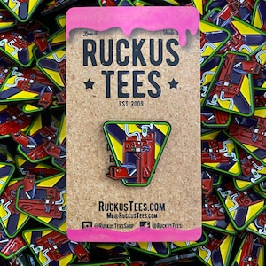 Könnte beinhalten: Ein dreieckiger Emaille-Pin mit einem roten und blauen Maschinendesign mit gelben und grünen Akzenten. Der Pin befindet sich auf einer Korktafel mit dem Text "RUCKUS TEES EST. 2009". Der Hintergrund ist mit ähnlichen Pins gefüllt.