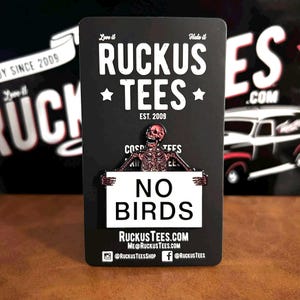 May include: A black rectangular pin featuring a skeleton holding a sign that reads "NO BIRDS." The pin is from Ruckus Tees, with the company name and website address printed on the bottom. The pin has a vintage aesthetic.