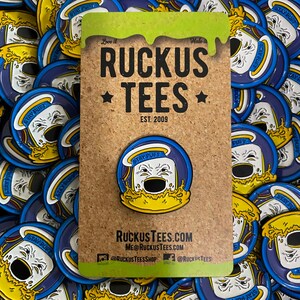 May include: A pin featuring a cartoon marshmallow character with the text "STAY PUFT" in a blue and yellow design. The pin is on a corkboard with the text "RUCKUS TEES EST. 2009". Additional pins are in the background.
