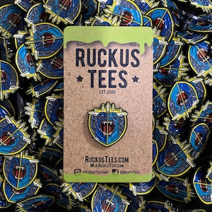 May include: A collection of enamel pins featuring a blue and yellow shield design with a snarling creature. The pins are on a corkboard card with the text "RUCKUS TEES" and the website address. The pins are scattered across a surface.