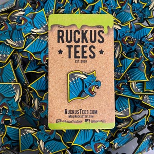 May include: A blue and yellow enamel pin featuring a stylized dragon design. The pin is on a corkboard with the text "RUCKUS TEES EST. 2009" and social media handles. Many similar pins are scattered around the corkboard.