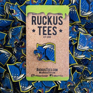 May include: A collection of blue and yellow enamel pins featuring a stylized dragon design. The pins are scattered around a corkboard card with the text "RUCKUS TEES EST. 2009" and the website address. The dragon pin is the focal point.