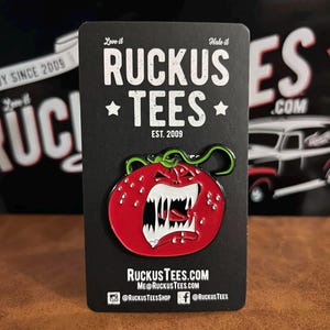 May include: A red enamel pin shaped like a strawberry with a menacing face, white teeth, and a green stem. The pin is on a black card with the text "Ruckus Tees" and "Est. 2009". The card also displays social media handles.