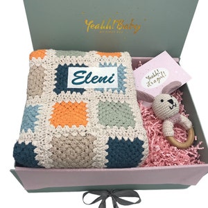 Op de afbeelding: Een cadeaudoos met een gehaakt dekentje, een gehaakte konijnenrammelaar en een kaart met de tekst "Yeah! It's a girl!" Het dekentje is veelkleurig en heeft een gepersonaliseerd naamschild met de tekst "Eleni".