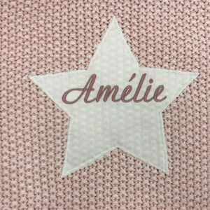 Peut inclure: Une couverture en tricot rose avec un patch en forme d'&eacute;toile blanche qui dit "Am&eacute;lie" en broderie rose.