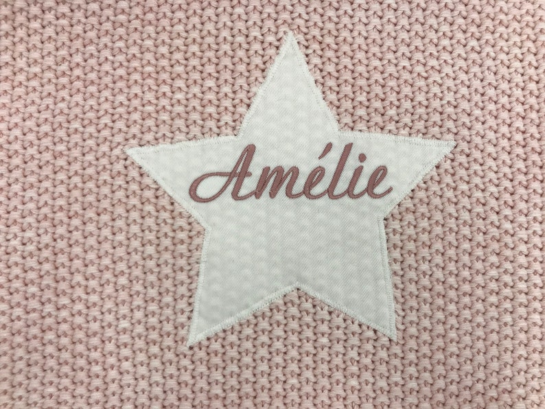 Peut inclure: Une couverture en tricot rose avec un patch en forme d'&eacute;toile blanche qui dit "Am&eacute;lie" en broderie rose.
