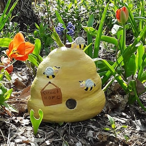 May include: A yellow, beehive-shaped birdhouse with cartoon bees and the words "Sweet Home" on a sign. The birdhouse is surrounded by green foliage and colorful flowers, including orange and red tulips.