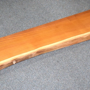 May include: A long, natural wood slab with a live edge. The wood has a warm, reddish-brown color and a smooth, polished finish.