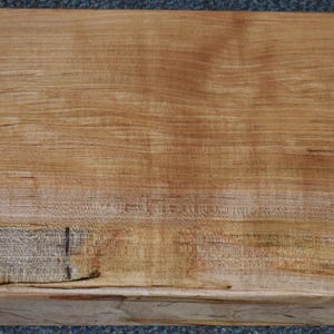 May include: A rectangular, unfinished wooden slab with a natural, light brown color and visible wood grain. The slab has a rough, textured surface and a dark spot near the center. The edges are raw and unfinished, showcasing the natural wood.