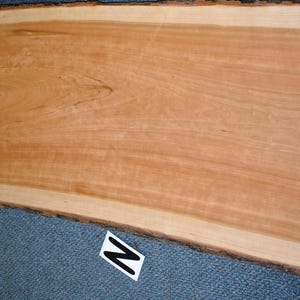 May include: A rectangular, unfinished wooden slab with a natural edge. The wood has a light brown color with darker grain patterns. The slab is likely for woodworking projects or as a decorative element.