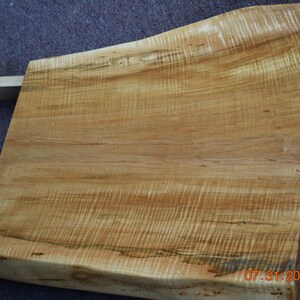 May include: Close-up of a wooden slab with a natural edge. The wood has a light brown color with darker grain patterns and a smooth surface. The date "07.31.2025" is visible on the bottom right corner.