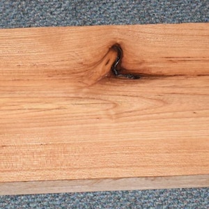 May include: A rectangular piece of unfinished wood with a natural, light brown color. The wood grain is visible, with dark brown knots and streaks adding visual interest. The wood appears to be ready for use in a woodworking project.