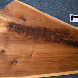 May include: A large, irregularly shaped wooden slab with a natural edge. The wood has a warm, reddish-brown hue with darker, swirling grain patterns. Several knots and imperfections add to the rustic aesthetic. A white sticker with a black L is in the upper right corner.