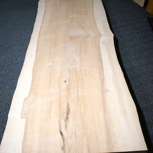 May include: A rectangular wooden slab featuring a natural live edge on both sides. The surface displays a light beige color with visible wood grain patterns, subtle knots, and variations in tone throughout the length of the board.