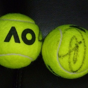 Puede incluir: Dos pelotas de tenis amarillas. Una pelota tiene las letras "AO" impresas en negro. La otra pelota tiene una firma negra.