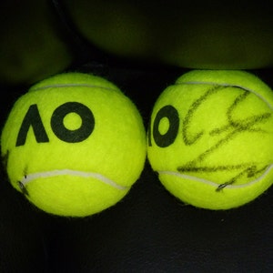 Puede incluir: Dos pelotas de tenis amarillas, una con las letras "AO" impresas y la otra con una firma negra.