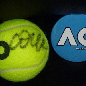 May include: A yellow tennis ball with the Australian Open logo and a black signature. A blue button with the Australian Open logo and the text "australian open" is next to the tennis ball.