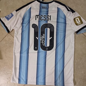 Op de afbeelding: Een wit en lichtblauw gestreept voetbalshirt met de naam MESSI en het nummer 10 op de achterkant. Het nummer 10 bevat een handtekening. Het shirt toont de tekst 1893, een FIFA World Cup 2026 patch en een FIFA World Cup embleem.