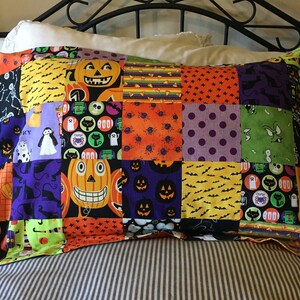 May include: A patchwork pillowcase with a Halloween theme. The pillowcase is made of various fabric squares featuring jack-o'-lanterns, bats, ghosts, and other spooky designs in orange, black, purple, and green. The pillowcase is rectangular.