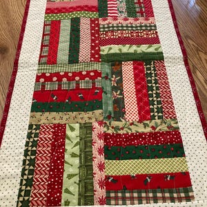May include: A rectangular quilted table runner featuring a patchwork design of vertical fabric strips in red, green, and white. The fabric patterns include holiday motifs like stockings, trees, and berries, framed by a white border with green dots.