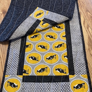 May include: A handmade table runner featuring a repeating pattern of black birds on yellow ovals against a gray background. The runner has a black border and a navy blue backing with a swirl design. The overall design is quilted.