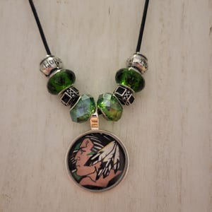 May include: A black beaded necklace with green glass beads and a silver charm featuring a Native American headdress. The charm has a black background and a colorful image of a person wearing a headdress. The beads on the necklace spell out the word "Hope".