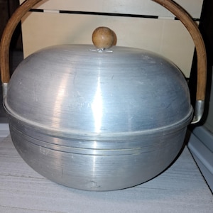 May include: A vintage aluminum cooking pot with a wooden handle and lid. The pot has a rounded shape and a wooden knob on top. The handle is arched and made of light brown wood. The pot is sitting on a light gray surface.