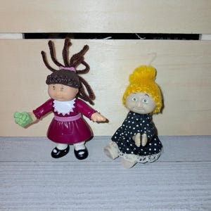 Può includere: Due bambole vintage Cabbage Patch Kids. Una bambola indossa un vestito bordeaux con colletto bianco e scarpe nere, che tiene in mano un oggetto verde. L'altra bambola indossa un vestito nero a pois bianchi e un berretto giallo lavorato a maglia.